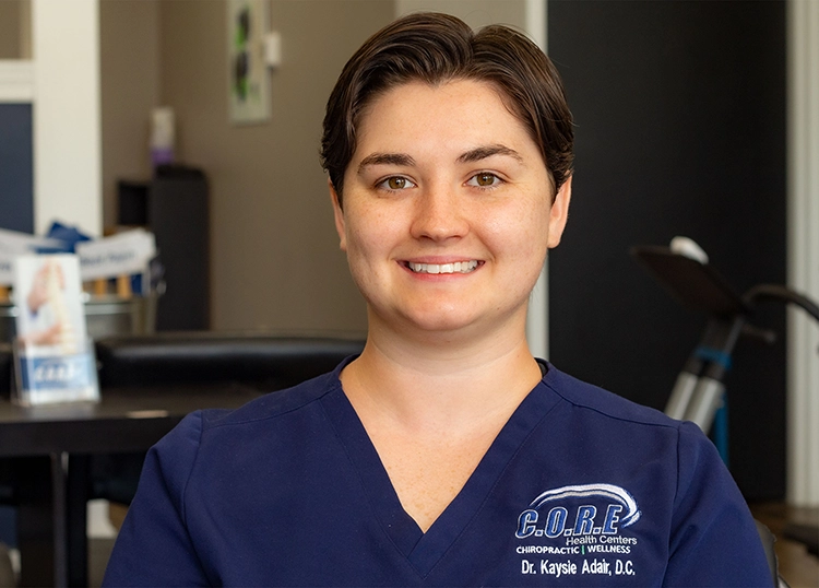 Smiling chiropractor in CORE Health Centers uniform, seated in a treatment room, representing Dr. Kaysie Adair, D.C.
