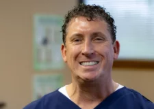 Dr-Grass-1080x1080-1-e1744647053739 (1) A man with short hair and glasses is smiling while looking at the camera. He is wearing a navy blue shirt over a white undershirt and seems to be in an indoor setting, possibly a chiropractic office, with posters and blurred objects in the background.