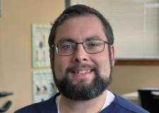 A man with short hair and glasses is smiling while looking at the camera. He is wearing a navy blue shirt over a white undershirt and seems to be in an indoor setting, possibly a chiropractic office, with posters and blurred objects in the background.