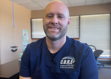 A man with short dark hair and a slight smile is wearing a blue scrub top with "C.O.R.E." and additional text embroidered on it. He appears to be in a chiropractic office, emphasizing pain relief through expert chiropractic care, with blurred background elements.
