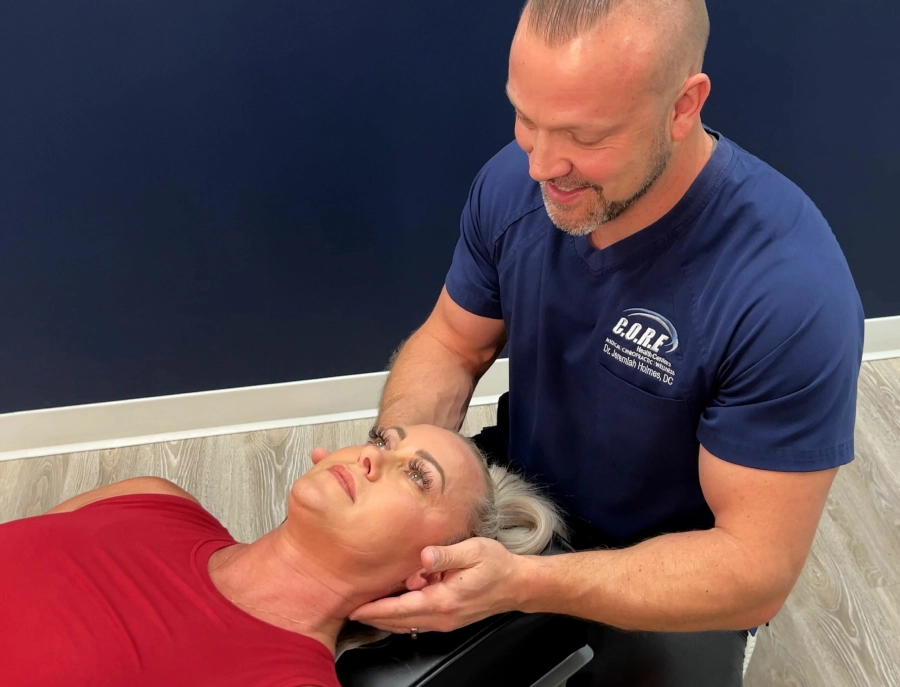 A chiropractor in a blue shirt is performing a chiropractic adjustment on a woman lying on a table. The woman, dressed in a red tank top, looks up while the chiropractor gently holds her head. They are in a health center with dark blue walls and wooden flooring.